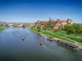 Widok na Wawel i Wisłę. Kilka łodzi pływa po rzece, a na brzegu zielony park i zabudowania.