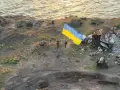 Na wyspie węży, pośród zniszczeń, żołnierze dumnie wznoszą ukraińską flagę. W tle morze i ruiny.