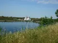 Biały dom z czerwonym dachem nad spokojnym pad rzeka, otoczony zielenią i wysoką trawą.