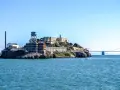 Słynne więzienie Alcatraz na skalistej wyspie, z charakterystyczną wieżą ciśnień i kominem, na tle błękitnego nieba i mostu.
