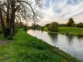 Spokojna rzeka na u, otoczona zielonymi brzegami i drzewami. W oddali widać ścieżkę i niebo z chmurami.