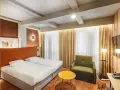 Przytulny pokój w hotelu Qubus Wrocław z dwoma łóżkami, zieloną sofą i żółtym stolikiem. Widoczne betonowe belki na suficie.