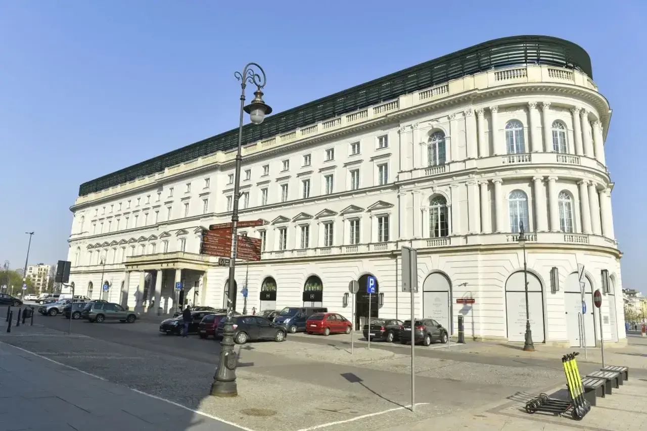 Elegancki budynek hotelowy, prawdopodobnie najdroższy hotel w Polsce, z białymi kolumnami i łukowymi oknami, stoi na tle błękitnego nieba.