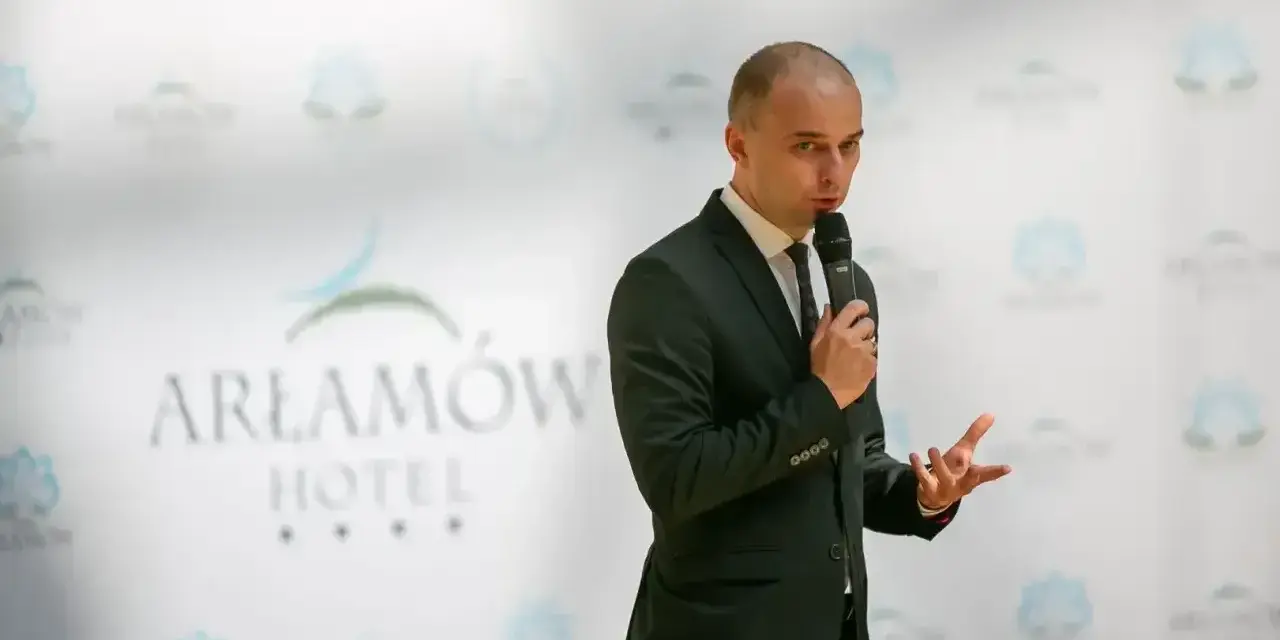 Mężczyzna w garniturze przemawia na tle logo Hotel Arłamów. Właściciel hotelu opowiada o swoich sukcesach.