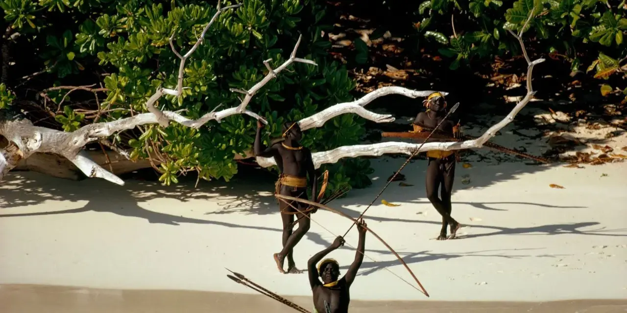 Trzech mężczyzn z plemienia Sentinelese, uzbrojonych w łuki i strzały, stoi na plaży. Sentinel Island, ich dom, chroni ich przed światem.