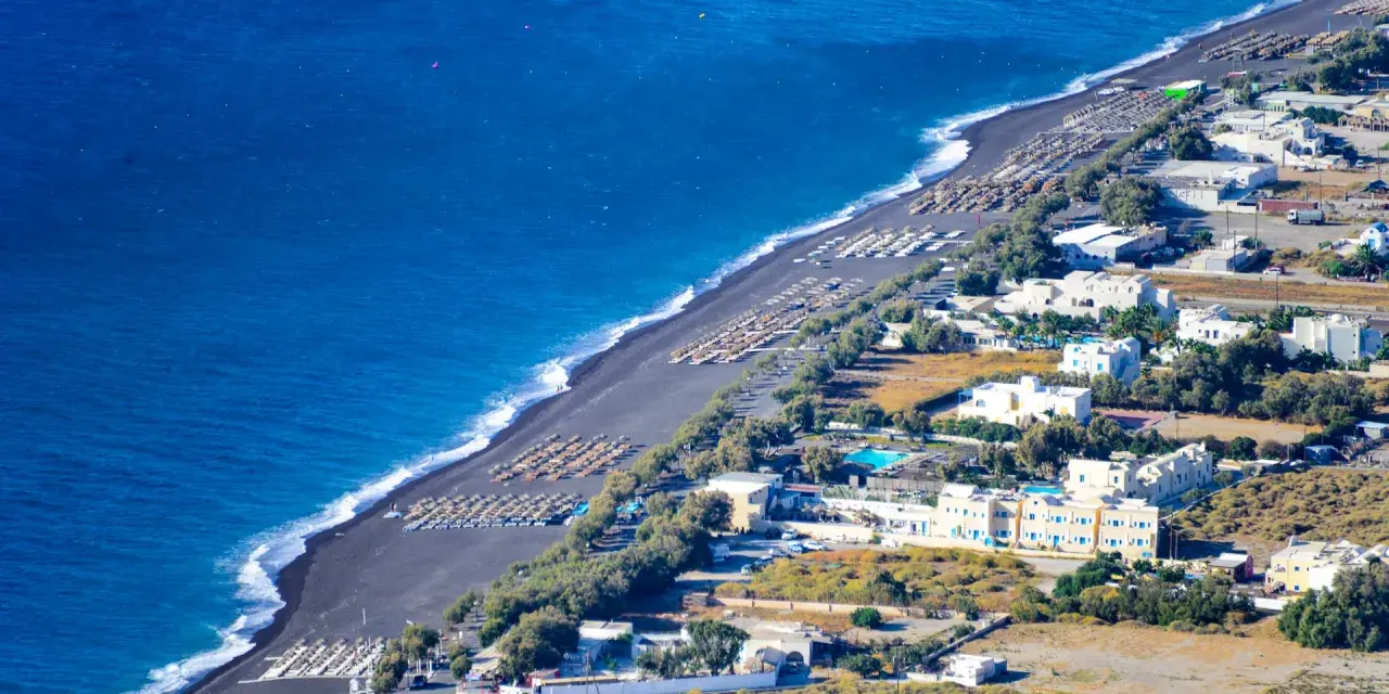 Santorini plaże z czarnym piaskiem, rzędy leżaków i białe budynki hotelowe nad turkusowym morzem.