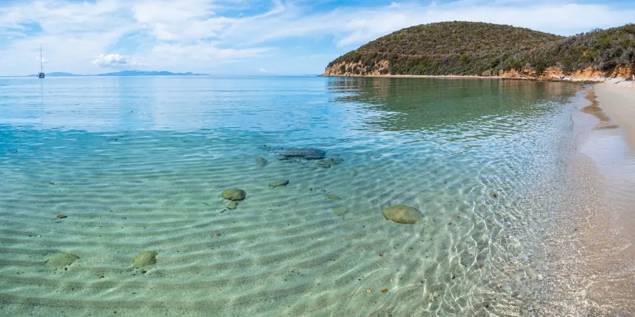 Piękne livorno plaże z krystalicznie czystą wodą i piaszczystym dnem. W oddali widać żaglówkę i zielone wzgórza.