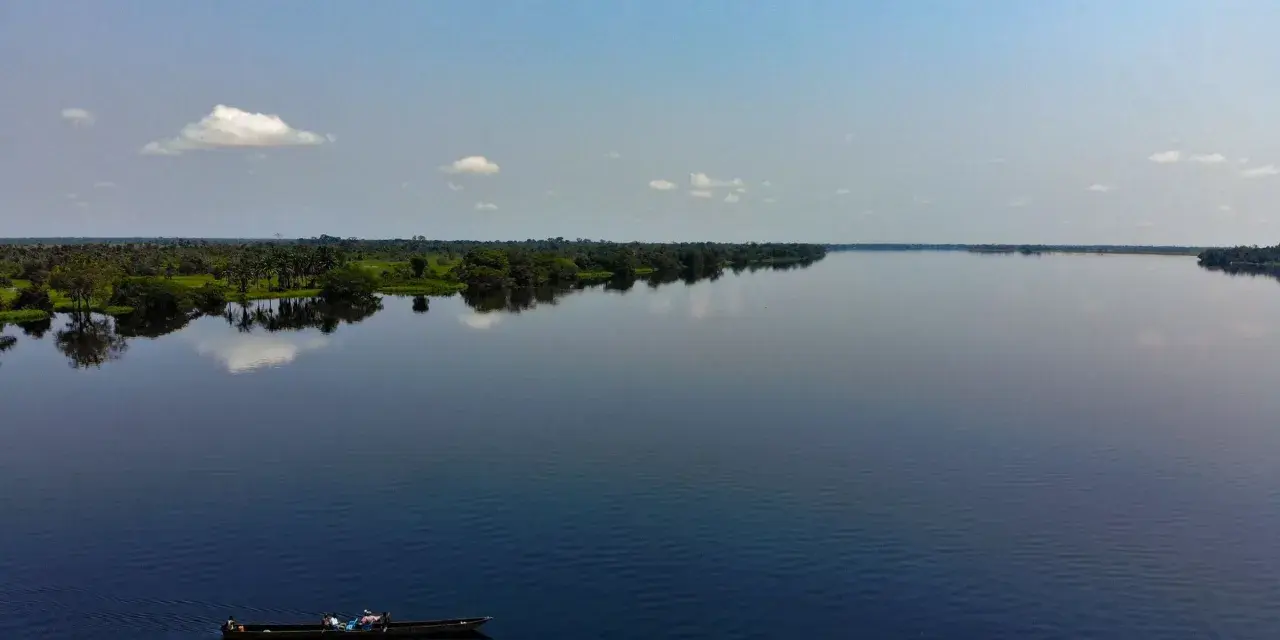 Szeroka, spokojna rzeka Kongo, odbijająca niebo i bujną zieleń brzegów. Na wodzie płynie łódź z kilkoma osobami.