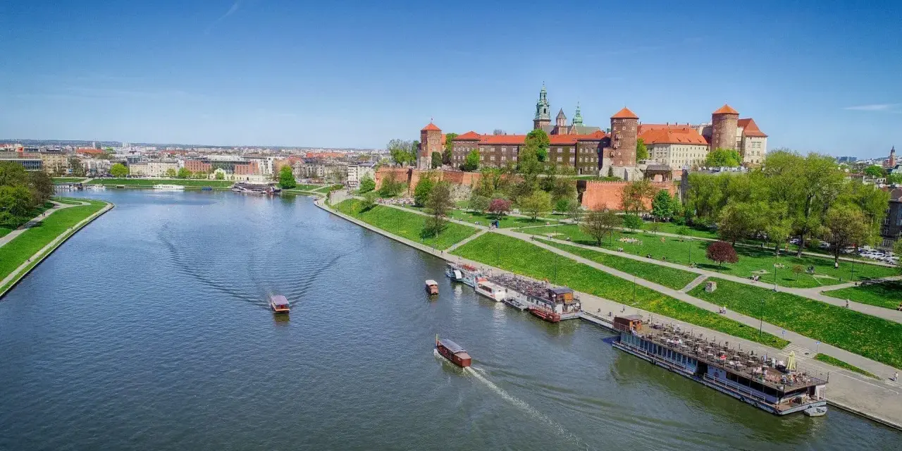 Widok na Wawel i Wisłę. Kilka łodzi pływa po rzece, a na brzegu zielony park i zabudowania.
