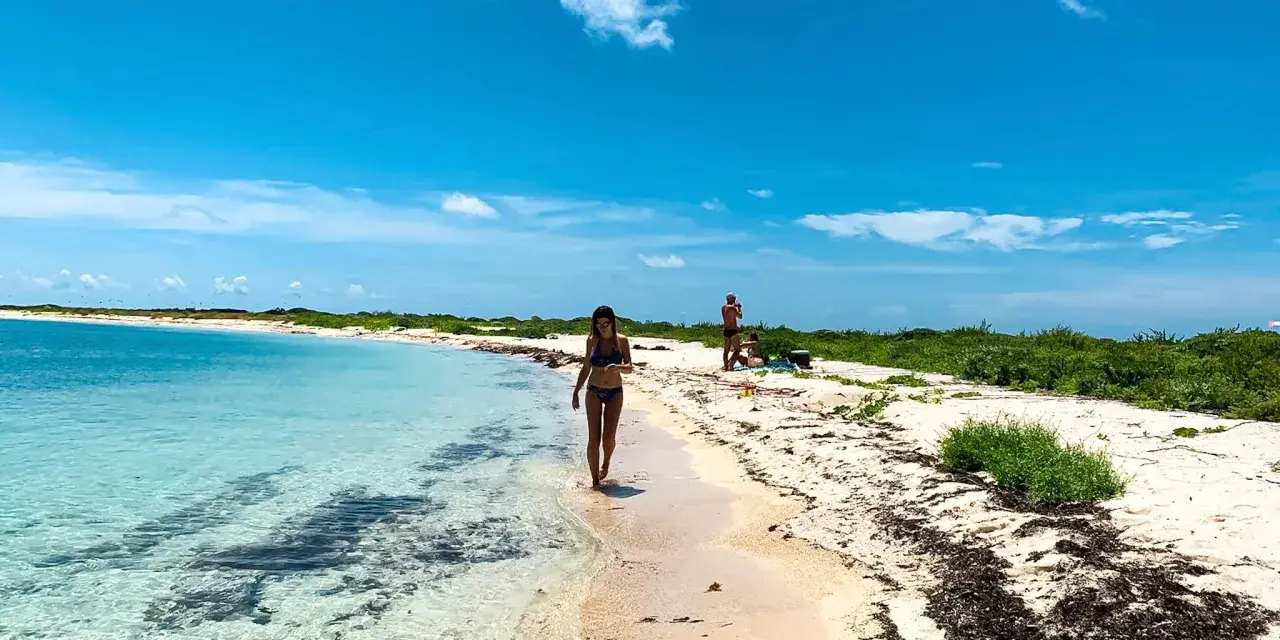 Kobieta spaceruje po piaszczystej plaży nad turkusową wodą. W tle widać innych ludzi i bujną zieleń. To idealne miejsce na wypoczynek nad Zatoką Meksykańską.
