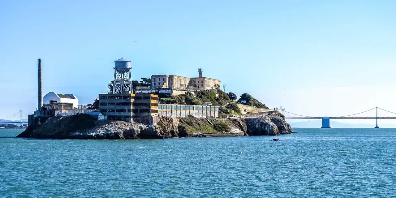 Słynne więzienie Alcatraz na skalistej wyspie, z charakterystyczną wieżą ciśnień i kominem, na tle błękitnego nieba i mostu.