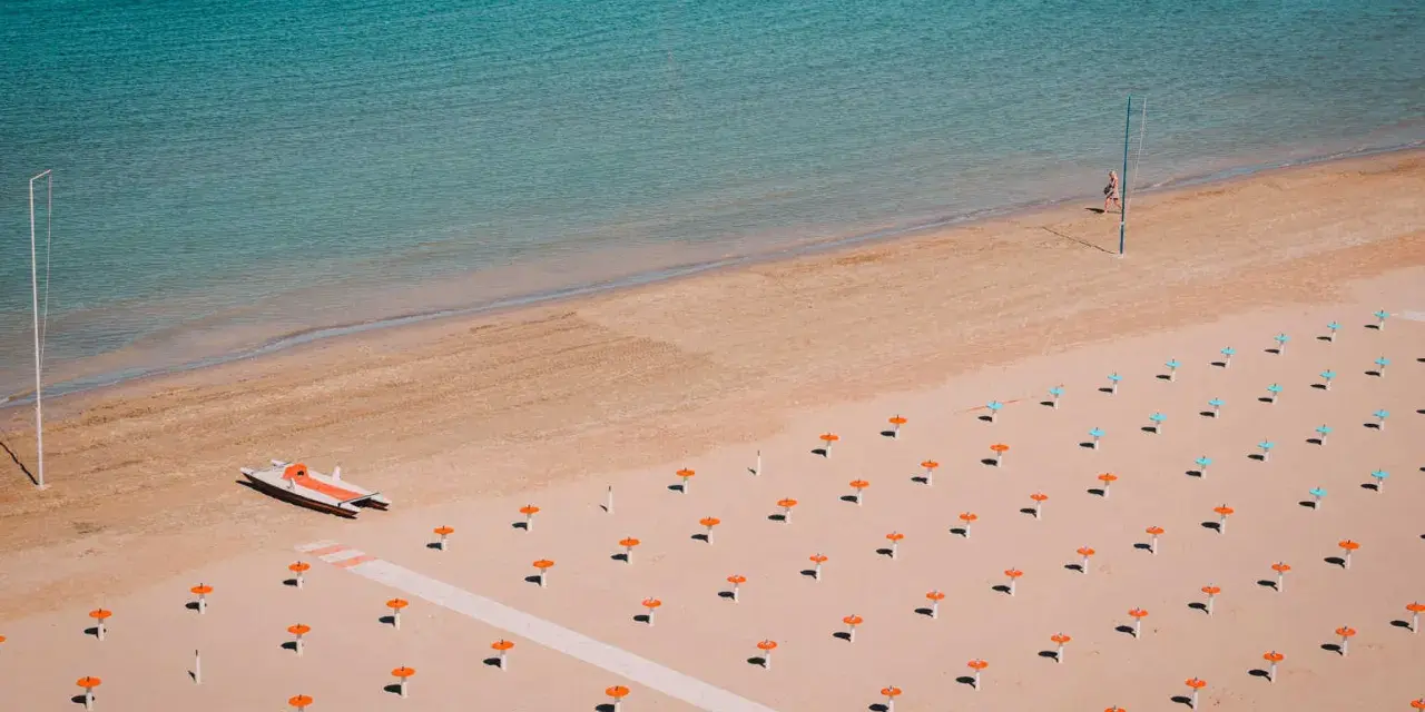 Pusta plaża w Rimini z rzędem pomarańczowych parasoli i turkusową wodą. Na piasku stoi mała łódź.