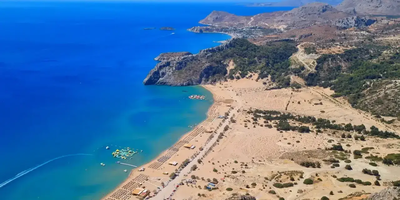 Malownicze **Rodos plaże** z turkusową wodą, piaszczystym brzegiem i zielonymi wzgórzami w tle. Widać wodny plac zabaw.