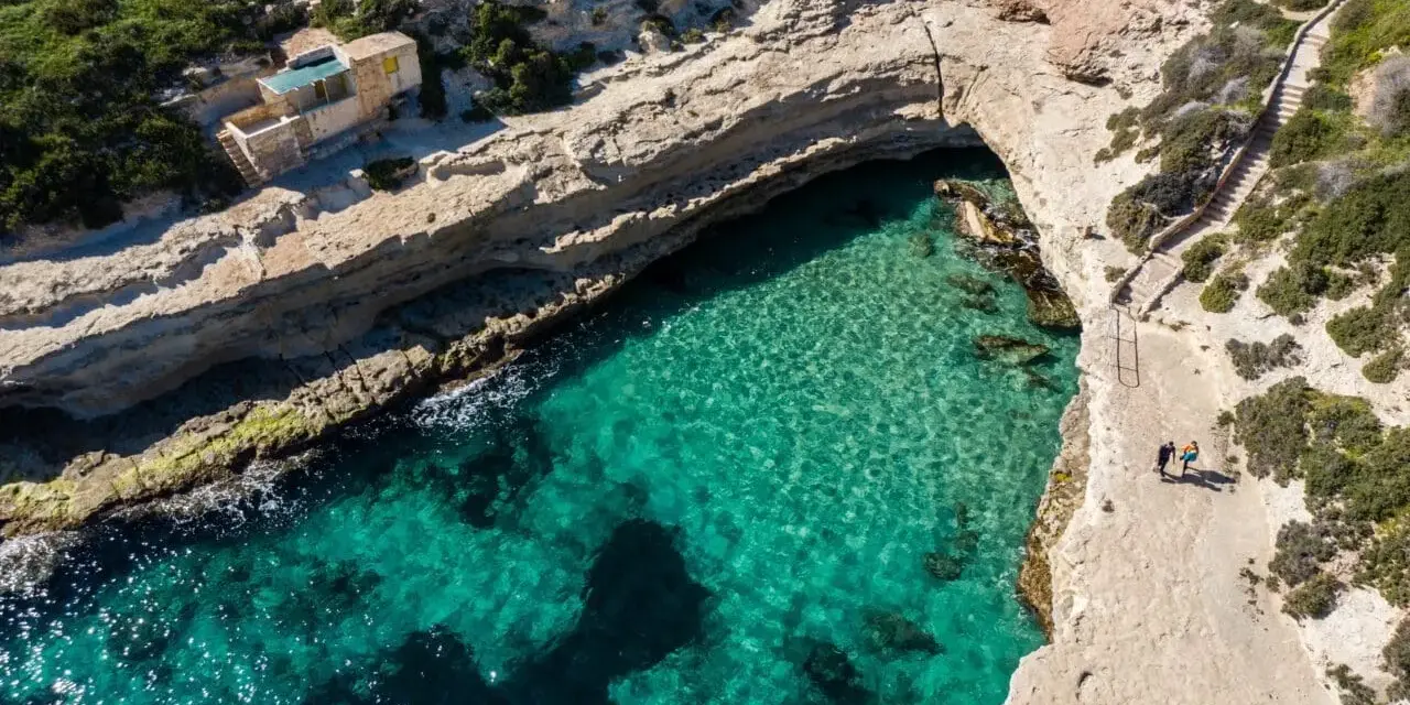 Malta plaże: turkusowa woda w zatoce, skaliste wybrzeże i mały domek. Dwie osoby spacerują ścieżką.