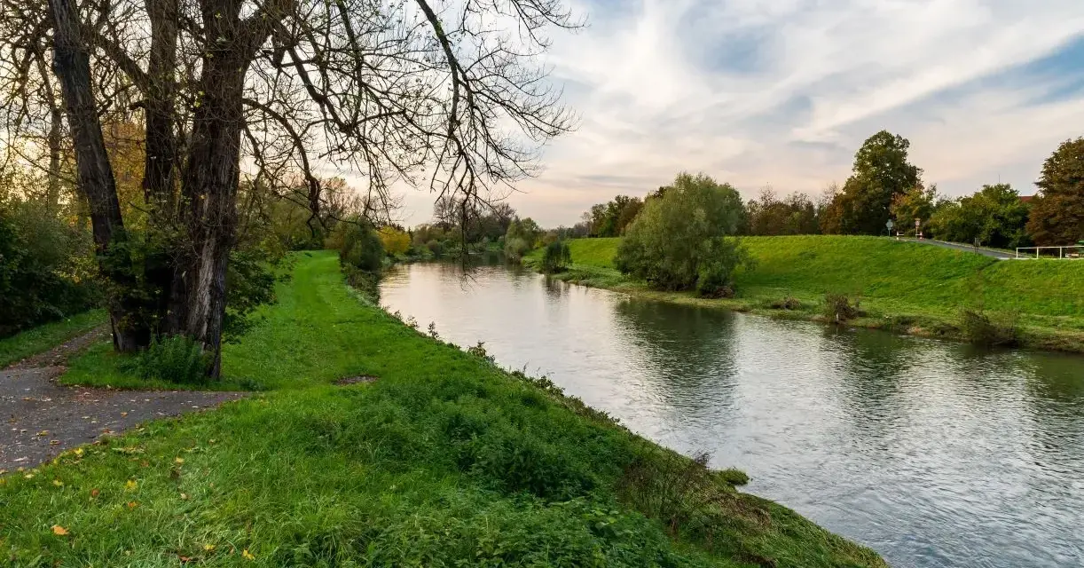 Spokojna rzeka na u, otoczona zielonymi brzegami i drzewami. W oddali widać ścieżkę i niebo z chmurami.