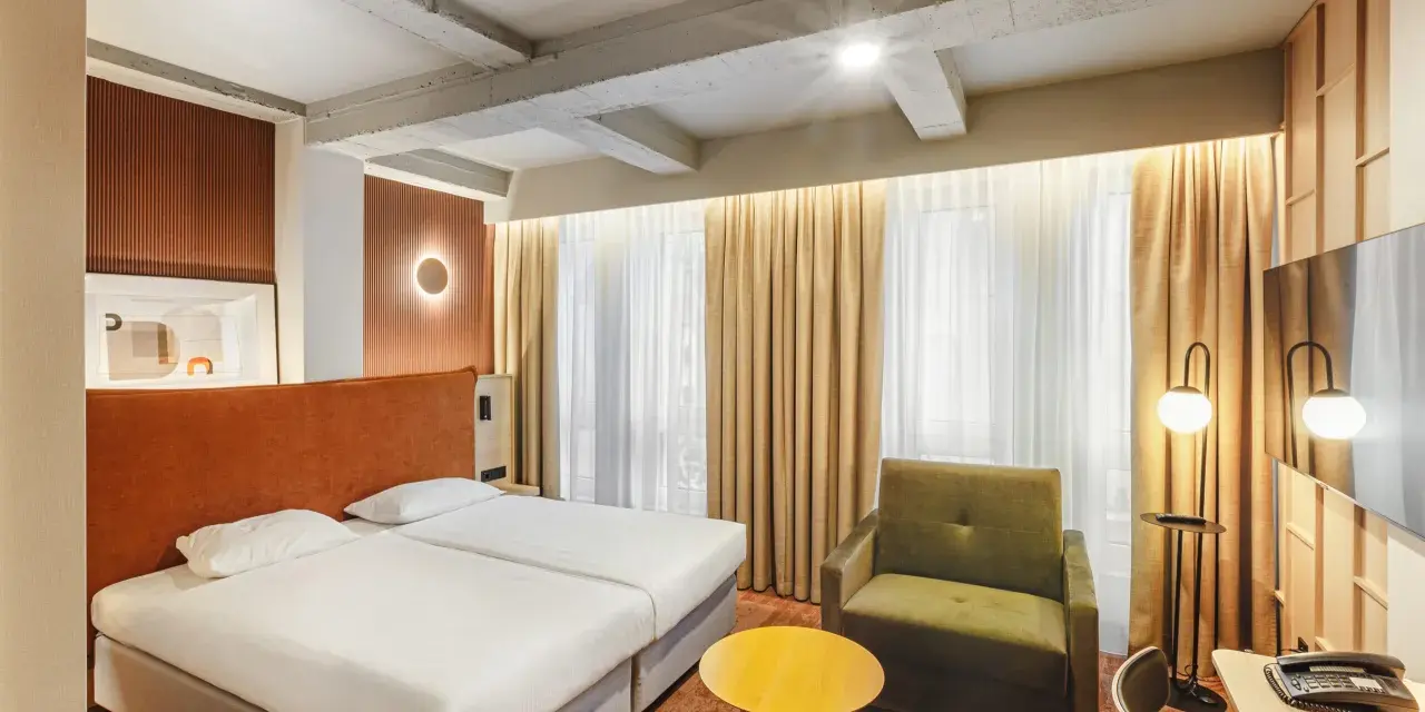Przytulny pokój w hotelu Qubus Wrocław z dwoma łóżkami, zieloną sofą i żółtym stolikiem. Widoczne betonowe belki na suficie.