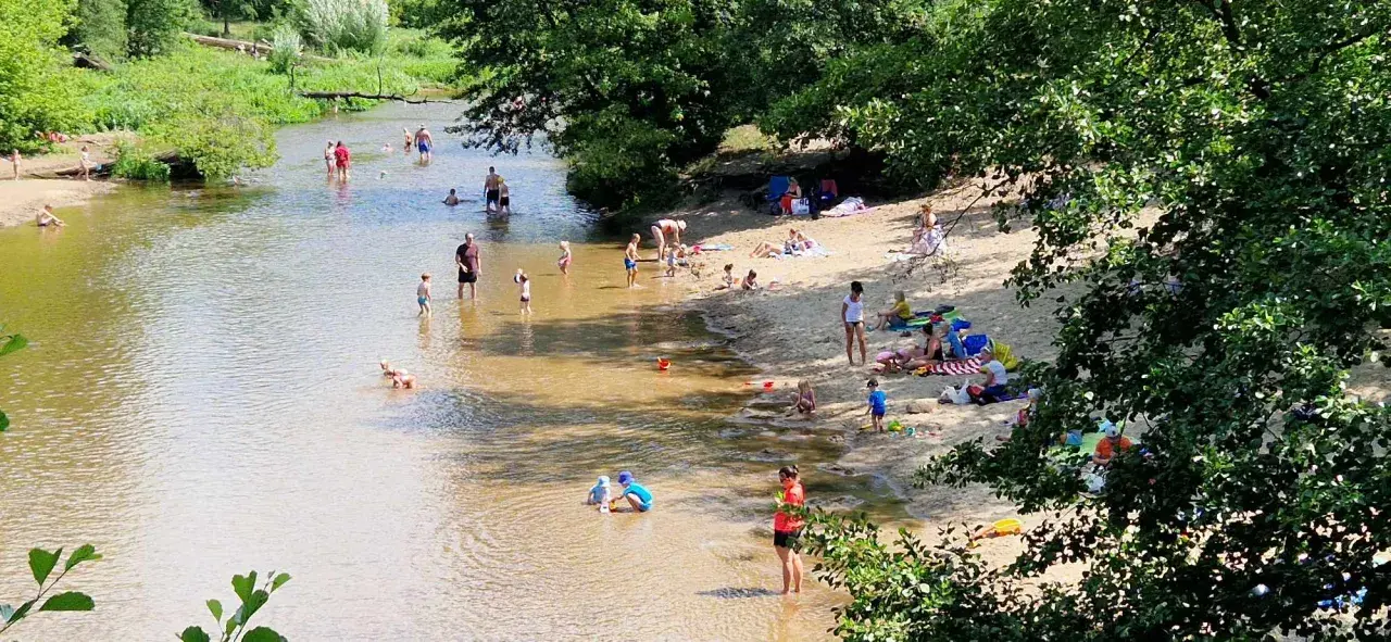Słoneczny dzień nad Świdrem. Ludzie odpoczywają na piaszczystej plaży i kąpią się w rzece. Dzieci bawią się w wodzie.