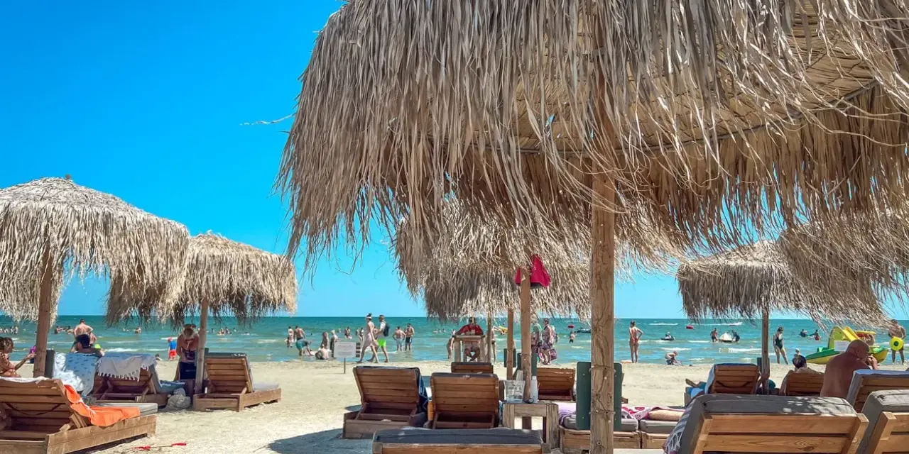 Słoneczne rumuńskie wakacje nad morzem. Leżaki i parasole z trawy na piaszczystej plaży, w tle ludzie kąpiący się w morzu.