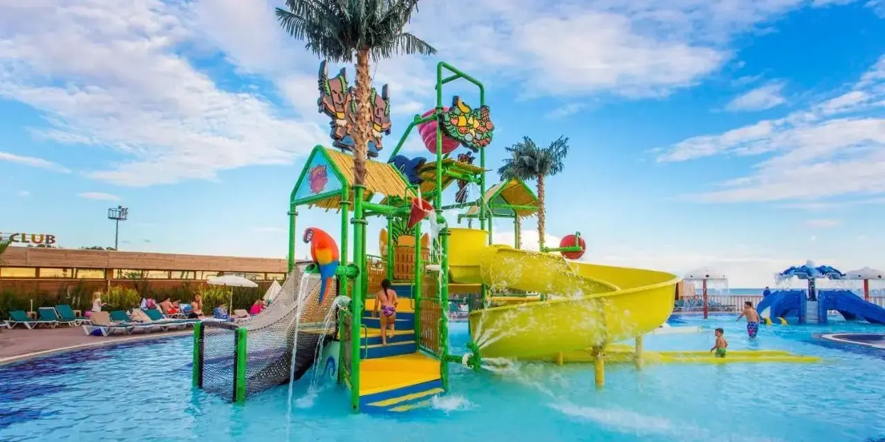 Kolorowy aquapark przy hotelu z palmami i zjeżdżalniami. Dzieci bawią się w wodzie, a na leżakach odpoczywają goście.