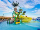 Kolorowy aquapark przy hotelu z palmami i zjeżdżalniami. Dzieci bawią się w wodzie, a na leżakach odpoczywają goście.