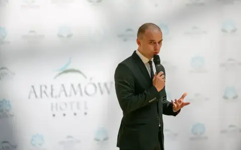 Mężczyzna w garniturze przemawia na tle logo Hotel Arłamów. Właściciel hotelu opowiada o swoich sukcesach.