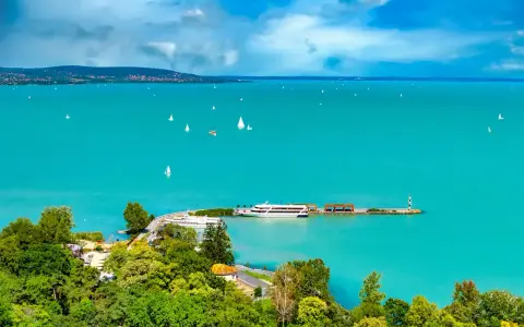 Turkusowe wody jeziora Balaton, pełne żaglówek i statków, z zielonymi wzgórzami w tle.