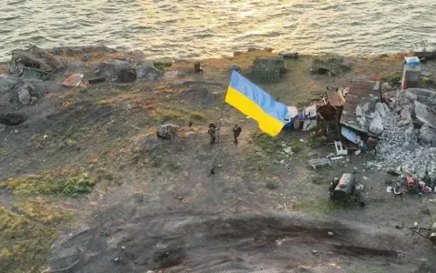 Na wyspie węży, pośród zniszczeń, żołnierze dumnie wznoszą ukraińską flagę. W tle morze i ruiny.