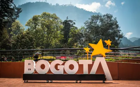 Napis BOGOTA z żółtymi gwiazdami, symbolizujący stolicę Kolumbii, na tle zielonych wzgórz i błękitnego nieba.
