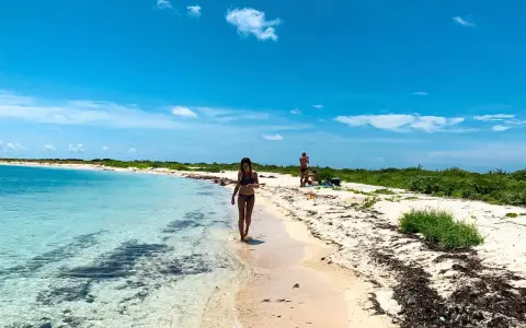 Kobieta spaceruje po piaszczystej plaży nad turkusową wodą. W tle widać innych ludzi i bujną zieleń. To idealne miejsce na wypoczynek nad Zatoką Meksykańską.