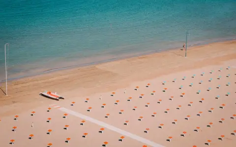 Pusta plaża w Rimini z rzędem pomarańczowych parasoli i turkusową wodą. Na piasku stoi mała łódź.