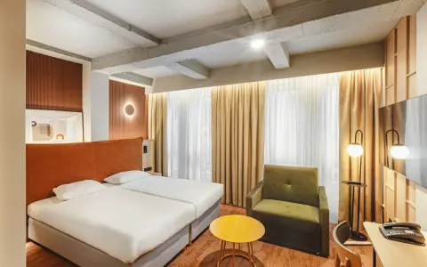 Przytulny pokój w hotelu Qubus Wrocław z dwoma łóżkami, zieloną sofą i żółtym stolikiem. Widoczne betonowe belki na suficie.