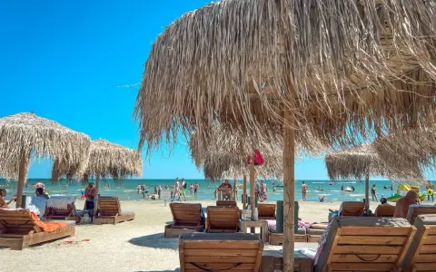 Słoneczne rumuńskie wakacje nad morzem. Leżaki i parasole z trawy na piaszczystej plaży, w tle ludzie kąpiący się w morzu.