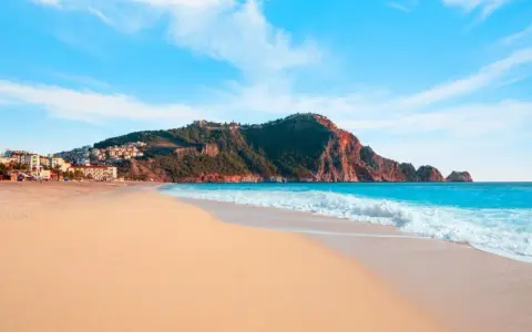 Plaża Kleopatry w Alanyi, z turkusowym morzem, piaszczystym brzegiem i górującym nad nią skalistym masywem z twierdzą.