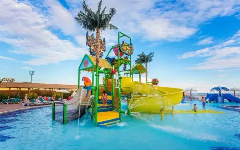 Kolorowy aquapark przy hotelu z palmami i zjeżdżalniami. Dzieci bawią się w wodzie, a na leżakach odpoczywają goście.