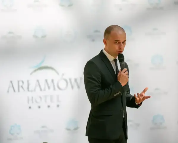 Mężczyzna w garniturze przemawia na tle logo Hotel Arłamów. Właściciel hotelu opowiada o swoich sukcesach.