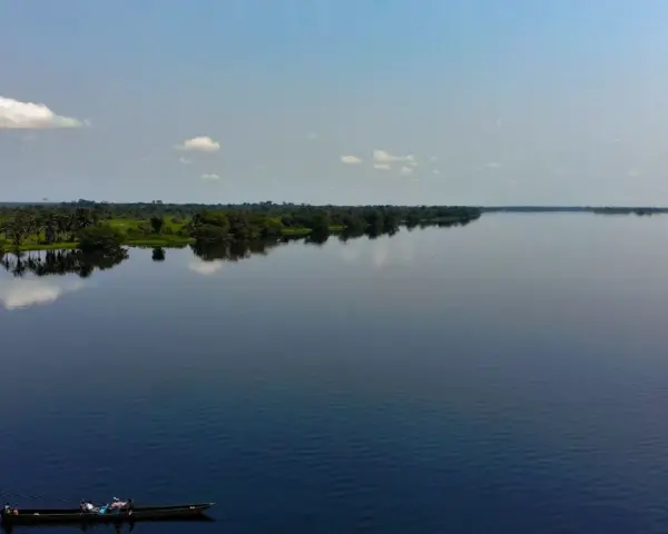 Szeroka, spokojna rzeka Kongo, odbijająca niebo i bujną zieleń brzegów. Na wodzie płynie łódź z kilkoma osobami.