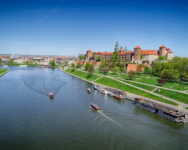 Widok na Wawel i Wisłę. Kilka łodzi pływa po rzece, a na brzegu zielony park i zabudowania.