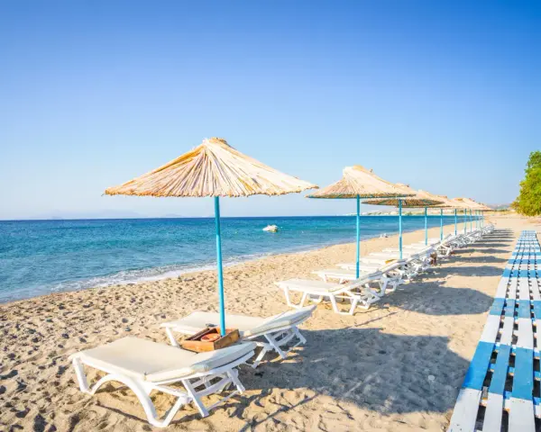 Plaża na wyspie Kos. Rzędy leżaków pod słomianymi parasolami, drewniana ścieżka i spokojne morze.