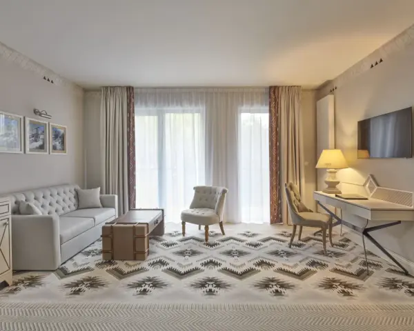 Elegancki pokój w hotelu 5 gwiazdkowym: sofa, stolik, fotele, biurko i TV. Wzory na dywanie i zasłonach dodają charakteru.