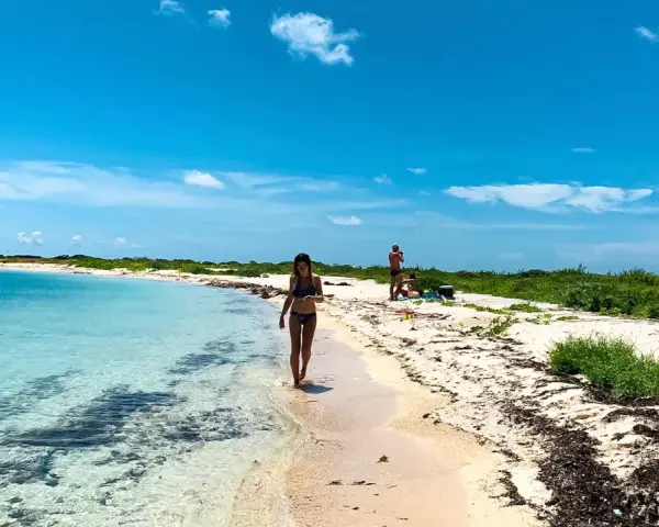 Kobieta spaceruje po piaszczystej plaży nad turkusową wodą. W tle widać innych ludzi i bujną zieleń. To idealne miejsce na wypoczynek nad Zatoką Meksykańską.