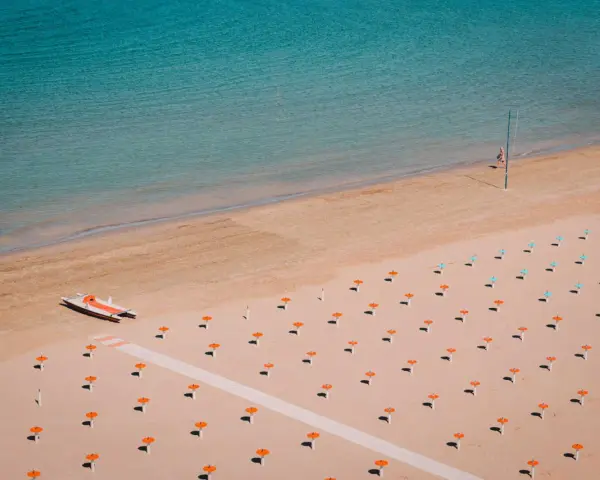 Pusta plaża w Rimini z rzędem pomarańczowych parasoli i turkusową wodą. Na piasku stoi mała łódź.