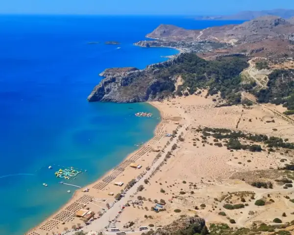 Malownicze **Rodos plaże** z turkusową wodą, piaszczystym brzegiem i zielonymi wzgórzami w tle. Widać wodny plac zabaw.