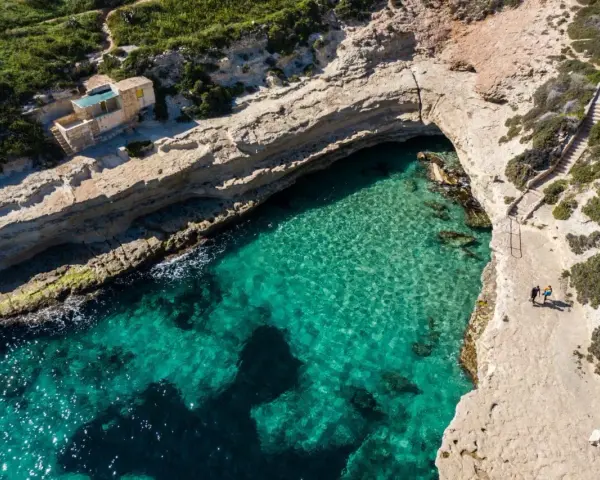 Malta plaże: turkusowa woda w zatoce, skaliste wybrzeże i mały domek. Dwie osoby spacerują ścieżką.