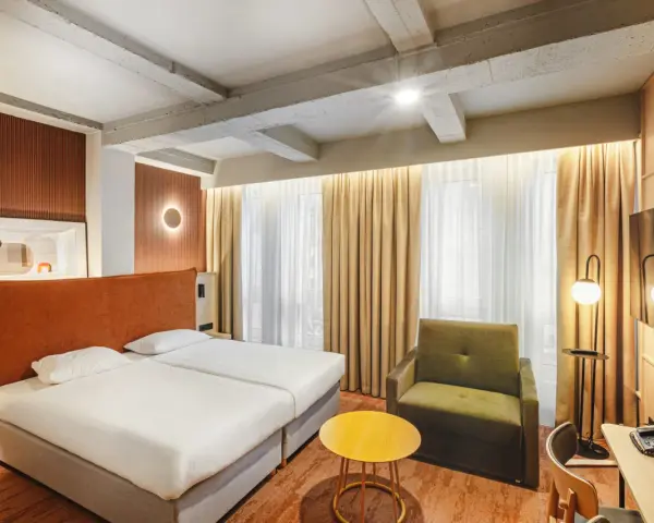 Przytulny pokój w hotelu Qubus Wrocław z dwoma łóżkami, zieloną sofą i żółtym stolikiem. Widoczne betonowe belki na suficie.