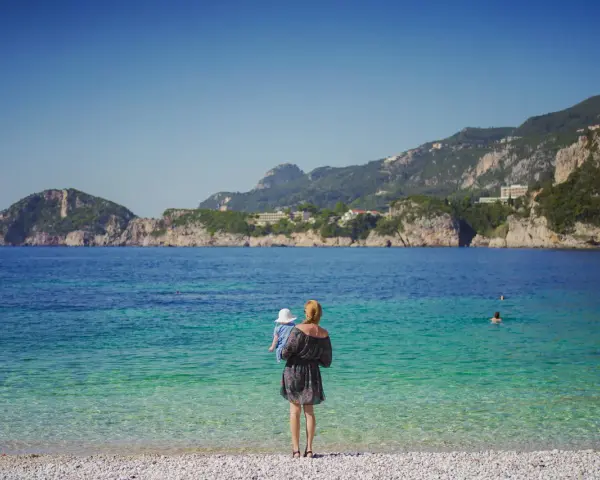 Kobieta z dzieckiem na rękach podziwia piękne Korfu plaże. Woda jest krystalicznie czysta, a w oddali widać góry.