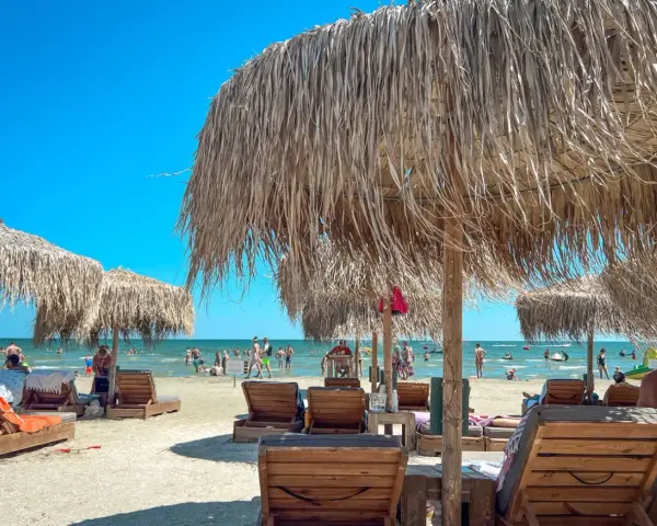 Słoneczne rumuńskie wakacje nad morzem. Leżaki i parasole z trawy na piaszczystej plaży, w tle ludzie kąpiący się w morzu.