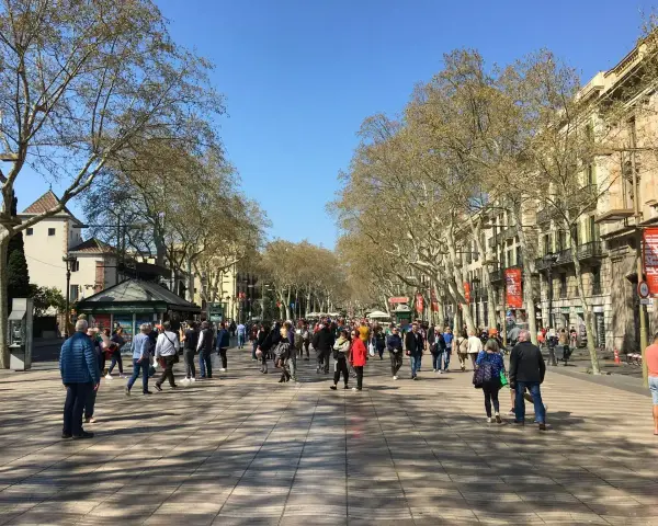 Tłum ludzi spaceruje po słonecznej La Rambla w Barcelonie, otoczonej drzewami i zabytkowymi budynkami.