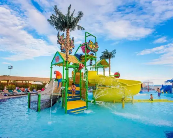 Kolorowy aquapark przy hotelu z palmami i zjeżdżalniami. Dzieci bawią się w wodzie, a na leżakach odpoczywają goście.
