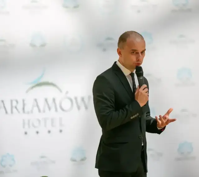 Mężczyzna w garniturze przemawia na tle logo Hotel Arłamów. Właściciel hotelu opowiada o swoich sukcesach.