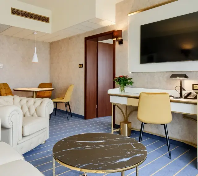 Elegancki pokój w hotelu Diament Wrocław: biała sofa, złote krzesła, biurko z telefonem i kwiatami, stolik kawowy.